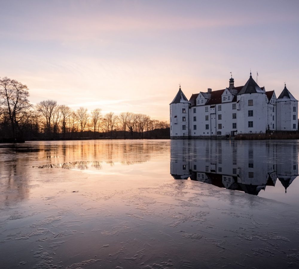 Das historische Wasserschloss in Glücksburg im Licht der untergehenden Wintersonne. Auf dem Schlossteich bildet sich eine Eisfläche